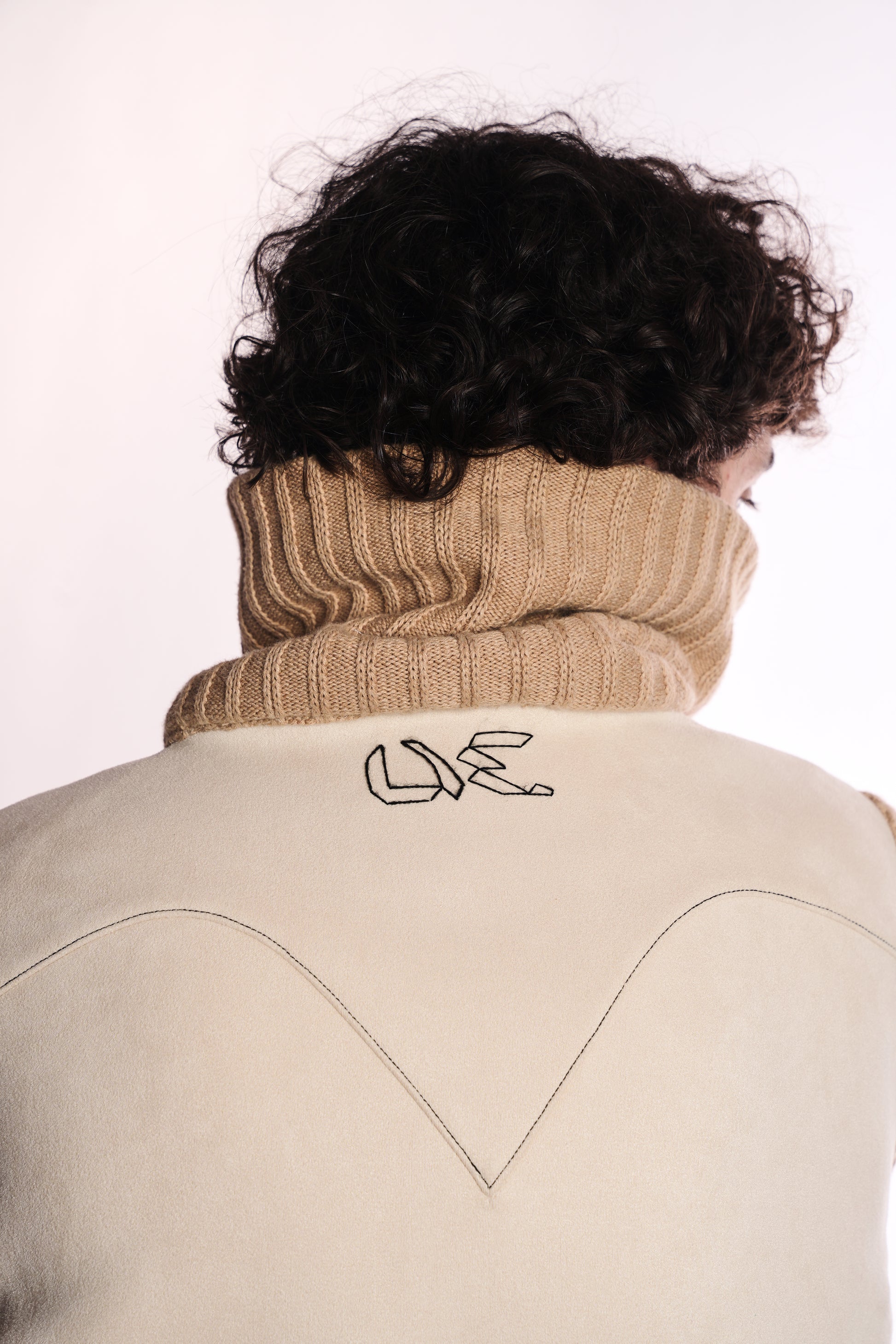 Person wearing a beige turtleneck sweater with a white jacket featuring a logo on a plain background